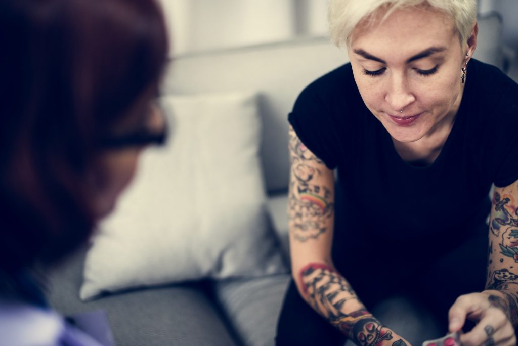A tattooed woman listens to a woman with glasses. This could represent how couples therapy and marriage counseling in Pasadena, CA can help repair relationships. Contact a marriage counselor to learn about infidelity counseling and other services.