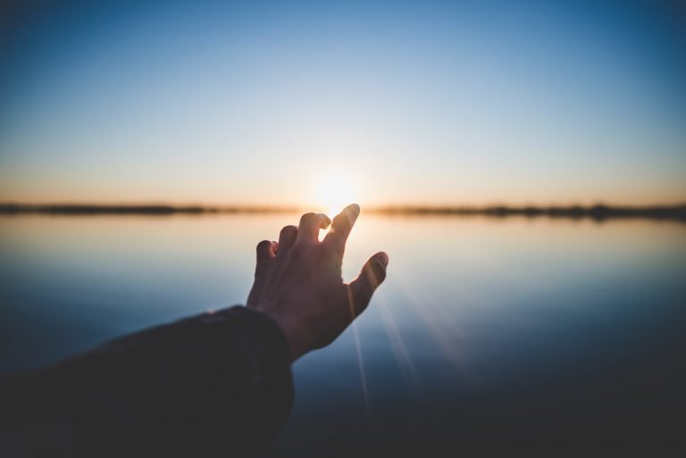 A hand reaches for the rising sun over a body of water. This could represent overcoming past trauma with a trauma therapist in Pasadena, CA. Contact us for trauma therapy in Sacramento, CA, PTSD treatment in Pasadena, CA, and other services. 95630 | 95688 | 95765