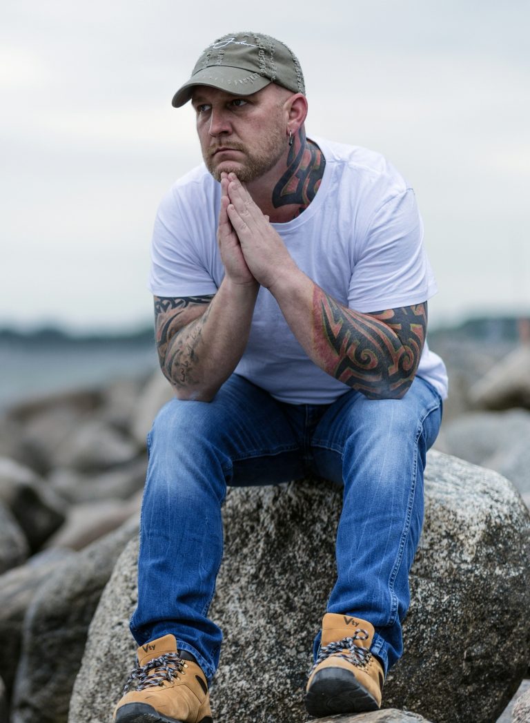 A tattooed man sits on a rock while appearing anxious. This could represent how anxiety symptoms can strike at any moment. Contact an anxiety therapist in Pasadena, CA for info on how panic attack treatment can support you. We also offer anxiety treatment in Sacramento, CA, and other services. | 95814 | 95688 | 95765