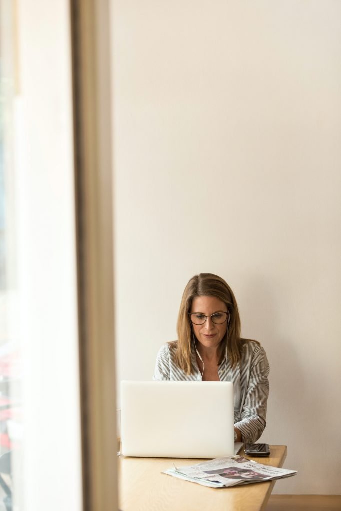 A woman sits at a desk against a blank wall as she types on her laptop. Contact an online therapist in California for support with online therapy wherever you are in the state!